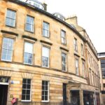 Cox & Co Estate agent in Edinburgh, Scotland. A curved, multi-story stone building with several large windows. The structure has an arched entrance on the lower left, metal railings on the terrace, and adjacent modern and historic buildings. The sky is bright, enhancing the light beige color of the stone facade.