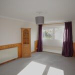 Cox & Co Estate agent in Edinburgh, Scotland. A spacious room with beige walls and wooden paneling on the lower half, situated in South Queensferry's EH30 area. The room features a large window with purple curtains, a radiator beneath it, and a wooden built-in corner shelf. The floor is carpeted in light grey for added comfort.