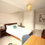 Cox & Co Estate agent in Edinburgh, Scotland. A neatly arranged bedroom with a double bed, white beddings, and a decorative cushion. It features a wooden headboard, bedside tables with lamps, a desk with a chair, and a large mirror. A wardrobe is built into the wall, and artwork decorates the space above a radiator. A small terrace offers castle-like views.