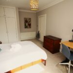 Cox & Co Estate agent in Edinburgh, Scotland. A bedroom with a bed covered in white linens and a teal pillow evokes the grace of a castle. Beside the bed is a small cabinet with a lamp. There's a desk with an office chair on the right, chest of drawers, and built-in cupboards on the left, with a painting and closed door in the background.