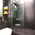 Cox & Co Estate agent in Edinburgh, Scotland. A modern bathroom with dark tiles features a glass-enclosed shower area. A wall-mounted toilet sits adjacent to a small sink. Wooden shelves holding toiletries and a toilet paper roll are mounted next to a mirror above the sink, reminiscent of the minimalistic charm you'd find in a castle terrace.