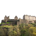 Cox & Co Estate agent in Edinburgh, Scotland. A historic castle sits atop a rocky hill, surrounded by lush greenery. The stone structure consists of multiple connected buildings with varying architectural styles, showcasing tall walls and small windows. A charming terrace offers a splendid view under the clear and bright sky in the background.