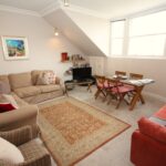 Cox & Co Estate agent in Edinburgh, Scotland. A cozy living room with beige and red sofas, an armchair, a TV on a stand, several shelves, and a small dining area with a wooden table and four chairs. The room features light-colored walls, a patterned rug, and a large window allowing natural light to flood in like the view from a castle terrace.