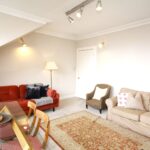 Cox & Co Estate agent in Edinburgh, Scotland. A cozy living room with three sofas arranged around a patterned area rug. One sofa is red, a second is beige, and a third is checkered. A dining table with chairs sits in the foreground. The white-walled room, reminiscent of a castle’s elegance, is illuminated by track lighting and a standing lamp.