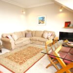 Cox & Co Estate agent in Edinburgh, Scotland. A bright living room features a beige sectional sofa with accent pillows, a plaid armchair, and a glass dining table with wooden chairs. Decorative elements include wall art, bookshelves, and a patterned area rug. There's a small TV on the right side, and doors opening to a charming terrace.