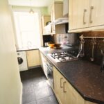 Cox & Co Estate agent in Edinburgh, Scotland. A narrow kitchen with light wood cabinets and black countertops. A gas stove, sink, and various kitchen appliances are visible. The window at the far end lets in natural light, revealing a distant terrace view, and a washing machine is also visible under the counter.