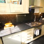Cox & Co Estate agent in Edinburgh, Scotland. A modern kitchen on Castle Terrace with light wood cabinets, black granite countertops, and a stainless steel gas stove. There is a dishwasher beneath the counter, a wooden cutting board with a bowl on top, a kettle, a toaster, and various utensils hanging on the wall.