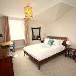 Cox & Co Estate agent in Edinburgh, Scotland. A cozy bedroom with a double bed adorned with white linens and decorative pillows, flanked by two small nightstands with lamps. A wooden desk with a chair is placed near a window with long curtains that open to reveal a charming terrace. A large mirror hangs on the wall above the bed, giving the room a stately, castle-like ambiance.