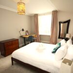 Cox & Co Estate agent in Edinburgh, Scotland. A spacious bedroom with beige walls, featuring a double bed with white linens and a teal pillow. A desk with a chair and lamp is placed under a window with beige curtains. A wooden chest of drawers and a large mirror complete the decor, hinting at castle elegance. A pendant light hangs from the ceiling.