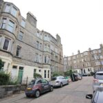 Cox & Co Estate agent in Edinburgh, Scotland. An image of Meadowbank Avenue shows a residential street lined with parked cars and stone apartment buildings. The buildings have multiple windows, some with bay windows, and a mix of green and beige doors. A few small trees and bushes are visible near the entrances. The sky is overcast.