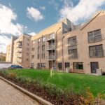 Cox & Co Estate agent in Edinburgh, Scotland. A modern apartment building featuring multiple floors with balconies on each level, located on Shrubhill Walk in Edinburgh (EH7). The facade is composed of beige bricks and large windows. In the foreground, a well-maintained lawn and shrubbery line a paved walkway, with several cars parked on the right side.