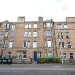 Cox & Co Estate agent in Edinburgh, Scotland. A mid-rise, beige stone apartment building with multiple windows in each unit. A single blue door serves as the entrance, and several black and green trash bins are lined up outside. Situated on Slateford Road, a paved road runs in front of the building.