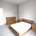 Cox & Co Estate agent in Edinburgh, Scotland. A minimalist bedroom on Slateford Road features light gray walls and carpet, a wooden bed frame with a white mattress, two matching wooden nightstands beside the bed, and a wooden dresser along the opposite wall. A white door is visible on the left side of the room.