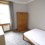 Cox & Co Estate agent in Edinburgh, Scotland. A minimalist bedroom on Slateford Road with a large window covered by gray curtains. The room features a wooden wardrobe, a matching chest of drawers, and a simple bed frame with a white mattress. The walls are painted light gray, and the carpet is a soft gray color.