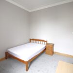 Cox & Co Estate agent in Edinburgh, Scotland. A small, minimalist bedroom off Slateford Road features light gray walls and a carpeted floor. The room includes a wooden single bed with a plain white mattress and a matching wooden bedside table with three drawers. A closed white door is visible on the adjacent wall.