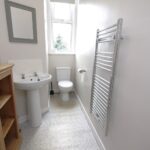 Cox & Co Estate agent in Edinburgh, Scotland. A small bathroom on Slateford Road features a pedestal sink, a toilet, and a wall-mounted towel warmer. A wooden shelving unit stands to the left of the sink, while a small window is situated above the toilet. A rectangular mirror hangs above the sink, and the floor is adorned with small mosaic tiles.