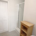 Cox & Co Estate agent in Edinburgh, Scotland. A bathroom on Slateford Road featuring a white door with a brass knob on the left, a glass-enclosed shower in the center, and a wooden shelf unit on the right. The floor is covered in small, light-colored tiles, and the walls are painted white.