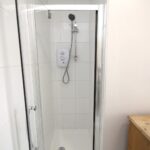 Cox & Co Estate agent in Edinburgh, Scotland. A small bathroom on Slateford Road features a tiled shower cubicle enclosed by a glass door. Inside the shower, there is a mounted showerhead with a handheld attachment. Adjacent to the shower is a wooden cabinet. The walls are plain and light-colored.