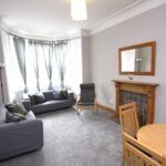 Cox & Co Estate agent in Edinburgh, Scotland. A modern living room on Slateford Road boasts a large window, gray walls, and carpet. It features a gray sofa, a wooden floor lamp, a wooden-framed mirror above a wooden fireplace, and a circular wooden dining table with four chairs. Light gray curtains cover the windows.