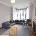 Cox & Co Estate agent in Edinburgh, Scotland. A cozy living room with gray carpeting and light gray walls. It features a large bay window with gray curtains, a gray sectional sofa with green cushions, a wooden armchair, a wooden fireplace with a mirror above, and a floor lamp. The ambiance is reminiscent of quaint homes on Slateford Road.