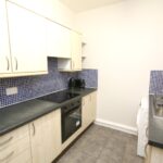 Cox & Co Estate agent in Edinburgh, Scotland. A compact kitchen on Slateford Road features light wood cabinets, a black countertop, a black electric stove and oven, and a stainless steel sink. The backsplash showcases small blue tiles. A toaster and kettle sit on the counter, while gray tiles cover the floor.