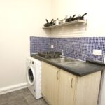 Cox & Co Estate agent in Edinburgh, Scotland. A compact kitchen corner on Slateford Road with light wood cabinets, a black countertop, and a stainless steel sink. Above the counter, there's a shelf holding pots and pans. A white washing machine is installed beneath the counter. Blue mosaic tiles form the backsplash.