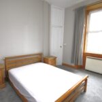 Cox & Co Estate agent in Edinburgh, Scotland. A minimalist bedroom on Slateford Road features a wooden bed frame with a white mattress, flanked by matching wooden bedside tables. A large window with gray curtains allows natural light to fill the room. The walls are light gray, and the floor is carpeted in a similar shade.